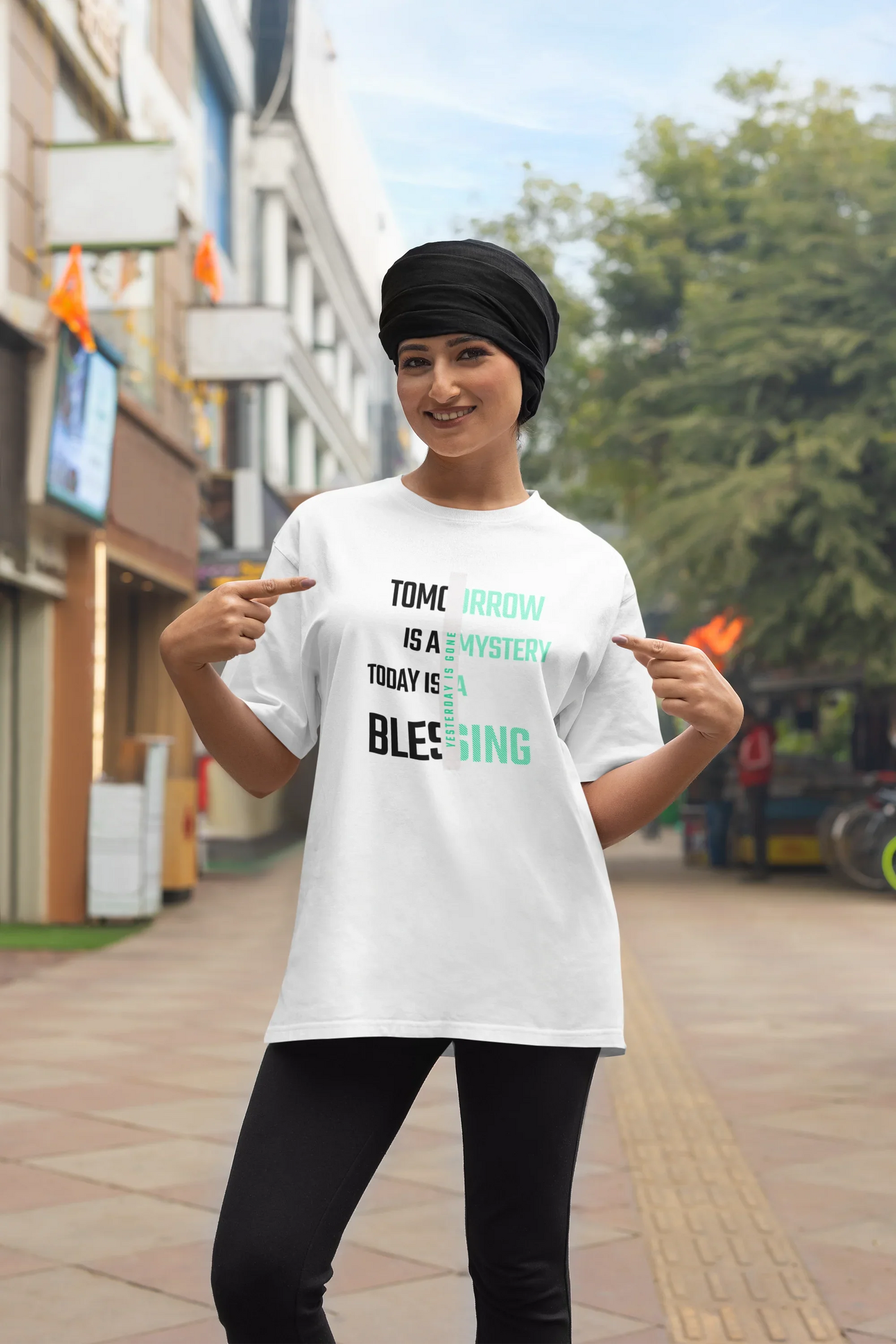 Smiling woman outdoors points at MY$TOAR luxury graphic tee, black turban, city street background