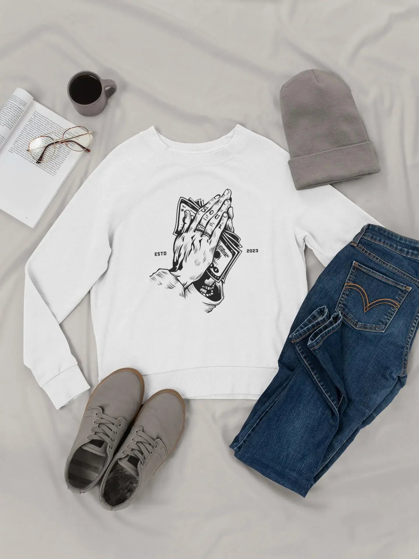 MY$TOAR luxury apparel flat lay with white sweatshirt featuring praying hands and money, blue jeans, gray beanie, shoes, and coffee.