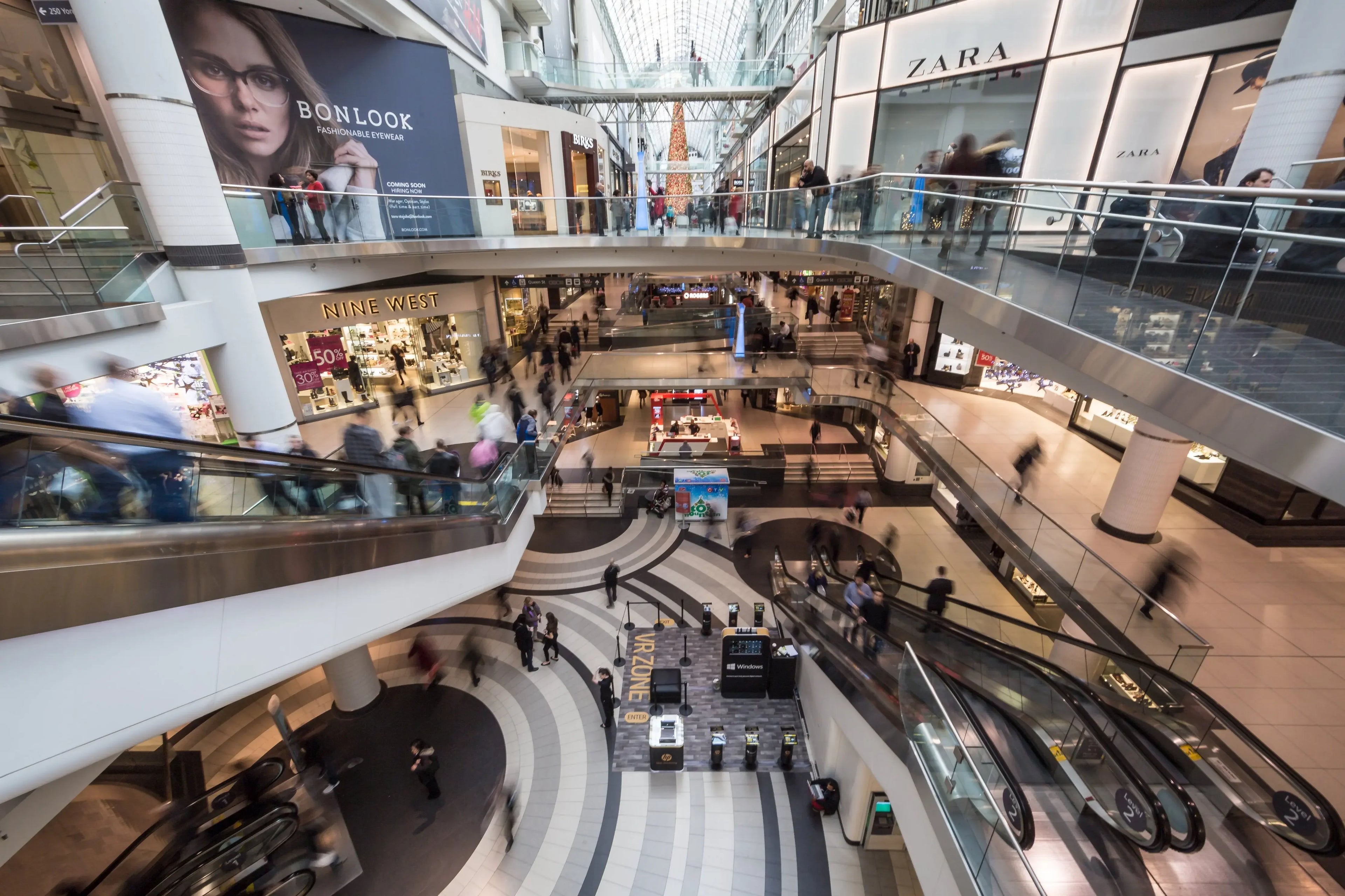 Busy modern shopping mall interior with escalators, retail stores, and people walking